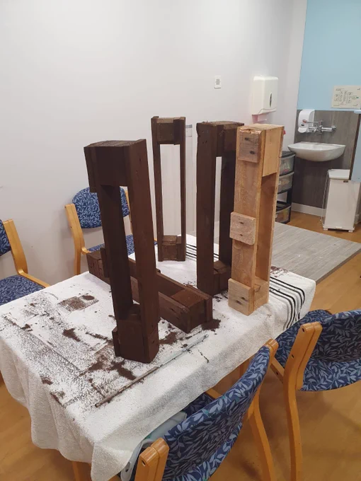 Wooden pallets arranged upright on a table covered with a white cloth and sawdust, in a room with chairs and a kitchenette in the background.