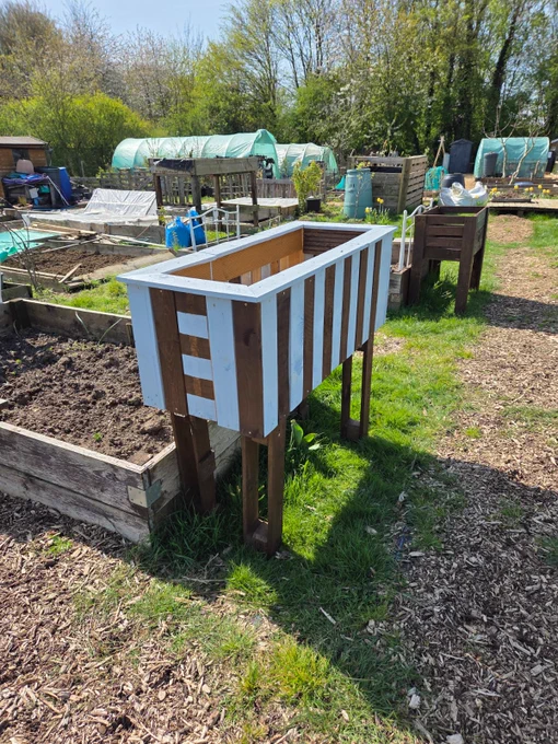 Raised garden bed in a community garden, featuring a wooden structure with white slats, surrounded by other gardening plots and equipment on a sunny day.