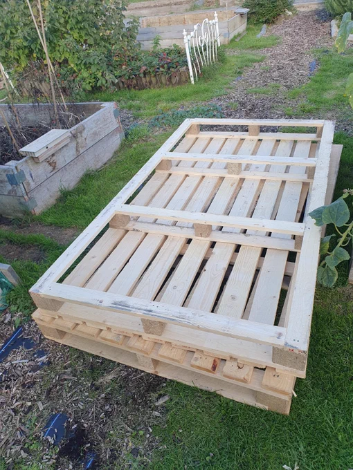 A stack of unused wooden pallets placed in a community garden with garden beds and greenery in the background.