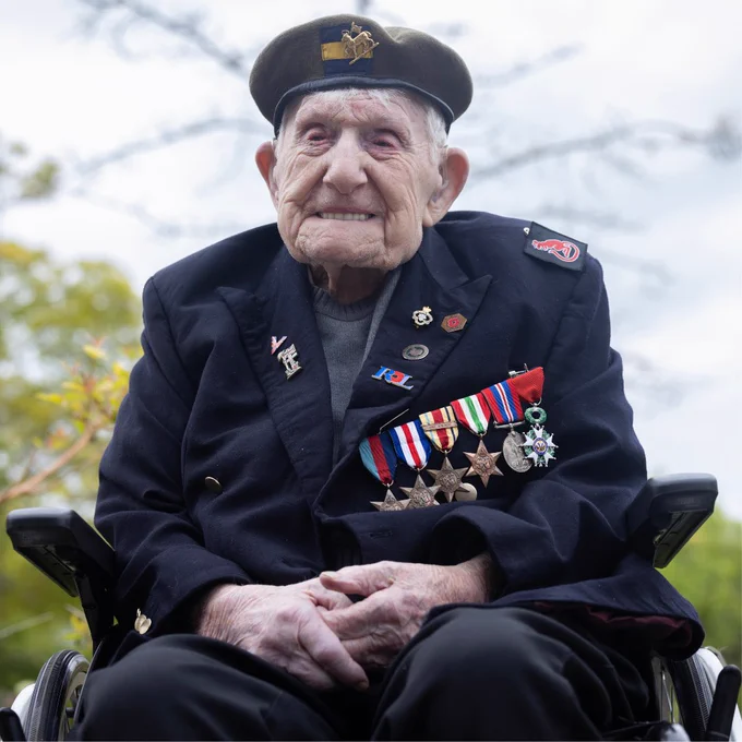 Donald Rose, wearing a military beret and a dark blazer adorned with numerous medals and pins, sits with his hands folded. His jacket displays service ribbons and honours, including the Legion d’Honneur. He has a gentle smile and is pictured outdoors at the National Memorial Arboretum against a soft, blurred background of trees and blue skies.