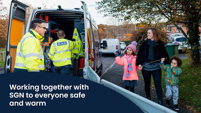 A happy family walking past two SGN engineers working at their van in a street. White text in a dark blue circle says: working together with SGN to keep everyone safe and warm.
