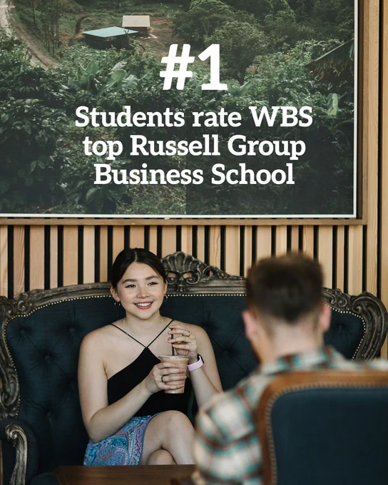 Two students are sat facing each other on ornate looking furniture beneath a large framed image hanging on the wall. White text overlaid reads "#1 Students rate WBS top Russell Group Business School".