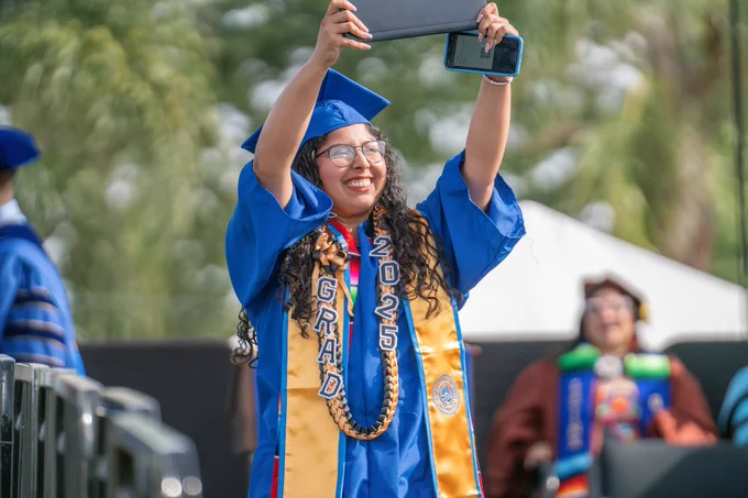Image of grad raising diploma above her head, walking down the ramp during commencement ceremony