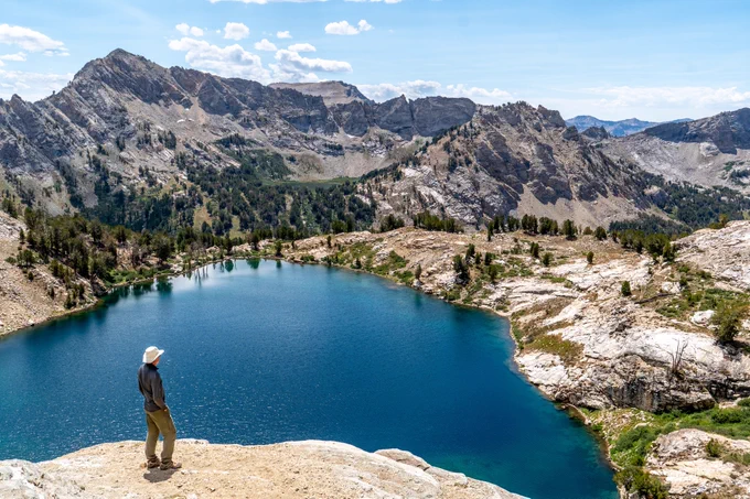 A hiker wearing a sun hat stands on a rocky ledge overlooking a deep blue alpine lake surrounded by pine trees and rugged mountain peaks under a bright blue sky with scattered clouds.