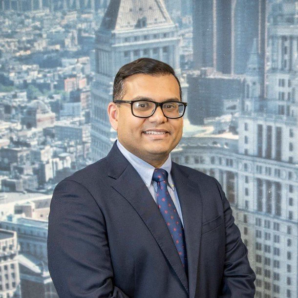 Hussain, wearing a dark suit and tie, poses in front of a photograph of the Dinkins building