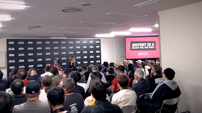 A somewhat low-quality screenshot of a video filmed over the heads of a smallish crowd in a convention panel room. The backdrop behind the panelists is adorned with a pattern of alternating "Star Wars" and "Celebration 2025" logos, and on the screen next to the panelists is a large title image: A background of a warm pink colour, on which is imposed a dark graphic shaped like duct tape, and the white title "History in a galaxy far, far away," subtitled in white: "Daughters of Ferrix with Chris Kempshall." The panelists are two people holding microphones, a man and a woman. The man has white skin and short hair, and is dressed in a red graphic tee shirt and an olive Imperial officer's hat. The woman is grinning, her microphone held up as she looks across the crowd: she has blonde choppy hair and bangs, the length of it draped over shoulders dressed in aw black leather jacket. 