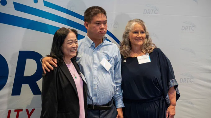 A man with his arm around a woman to his right and a woman standing to his left posing in front of a large DRTC backdrop.
