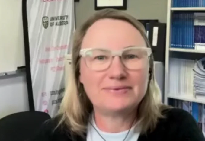 Person wearing glasses, smiling in an office setting with a University of Alberta banner in the background.