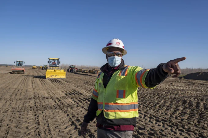 Man in safety gear in a field of bare soil points past the camera with bulldozers and other heavy equipment in the background. 