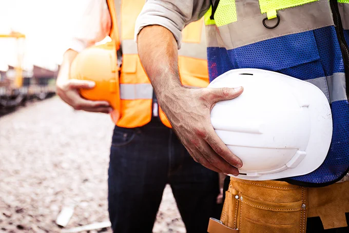Close-up of two construction workers holding safety helmets, wearing reflective vests and standing on a gravel site.