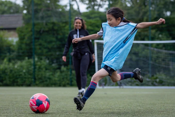A 9-year-old (approximately) medium-skinned girl dribbling a ball across a football pitch. She is wearing a bright blue bib. Her coach watches on the background, smiling widely.