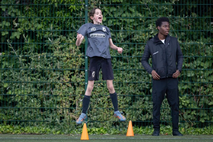 A light-skinned girl aged approximately 12 years old, in mid-air, mid-jump on the pitch sidelines. She is cheering on her teammates next to her dark-skinned coach.