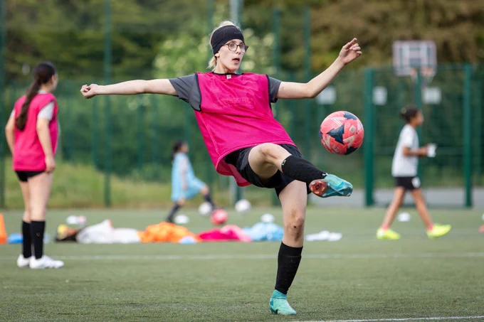 A light-skinned teenage girl kicking a red football up into the air. Her blonde hair is tied-up in a low ponytail, covered by a wide black sweatband.