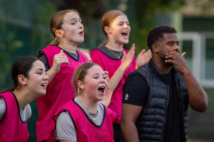 Four light-skinned girls aged 7-16 and dressed in red bibs, stand on the pitch sidelines cheering on their team mates with their dark-skinned coach.