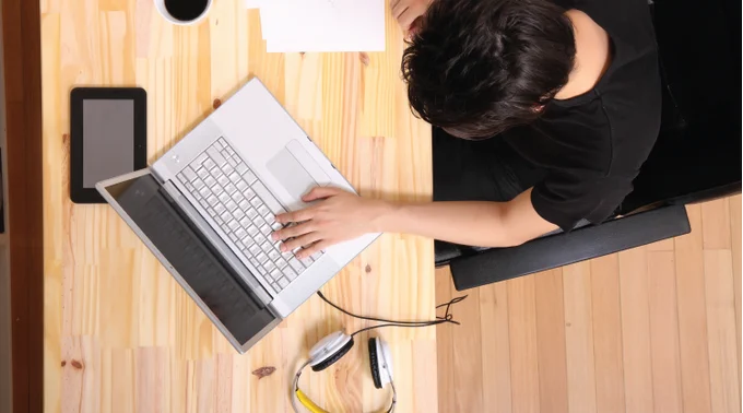 Overhead view of an individual working on a laptop at a wooden desk with scattered papers and a smartphone beside them.
