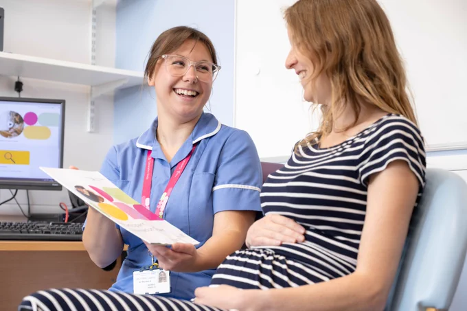 A clinical member of staff is sat next to a pregnant woman in a consultation room. She is showing her an information leaflet about the Generation Study.