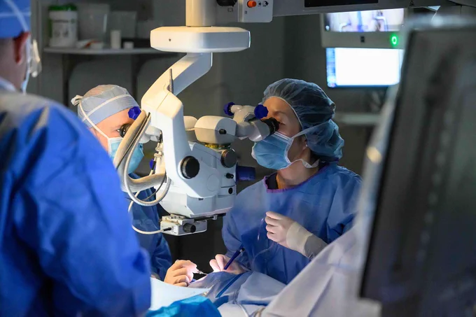 Surgery in progress in the OR, a doctor looking through a scope down at a patient while other medical professionals look on. 