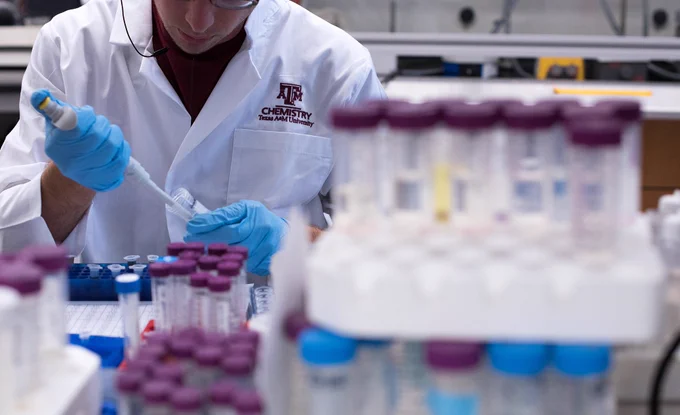 A person in a lab coat labeled "Texas A&M Chemistry" is pipetting a sample in a lab with numerous test tubes and vials in the foreground.