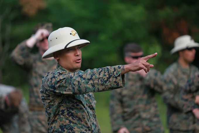 Marine instructor points forward while leading training outdoors, wearing camouflage uniform and pith helmet.