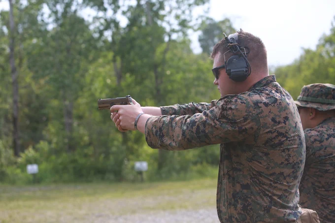 Marine aims pistol downrange during marksmanship training, wearing ear protection and sunglasses.