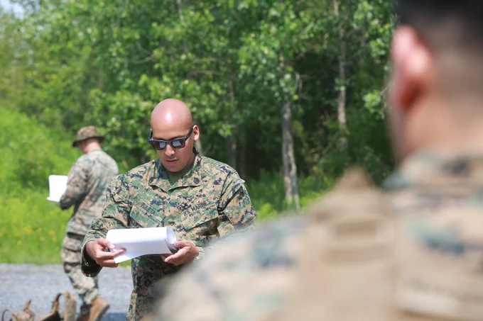 Marine in camouflage uniform reviews notes during field training with others visible in the background.