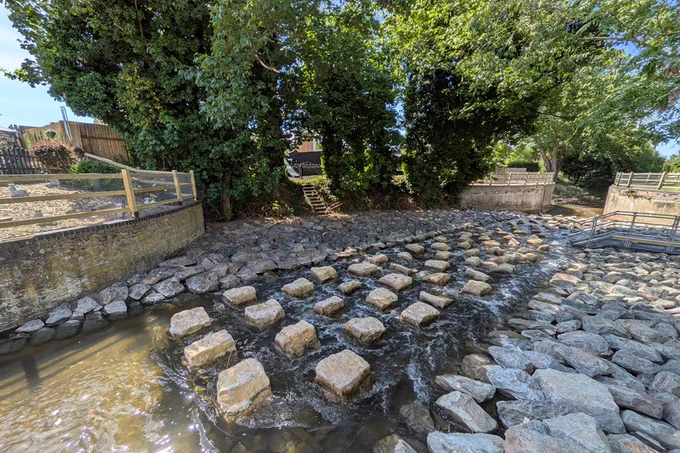The new fish pass across the River Lark with lush trees on the banks and a footbridge nearby on a sunny day.