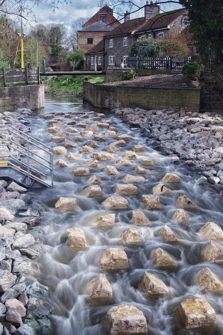 View of a new fish pass flowing through a village, with a metal stairway leading to the water's edge and traditional brick buildings in the background.