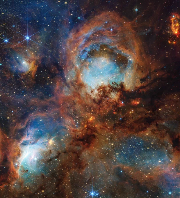 A section of the Cat’s Paw Nebula. Four roughly circular areas are toward the center of the frame: a small oval toward the top left, a large circle in the top center, and two ovals at bottom left and right. Each circular area has a luminous blue glow. Brown-orange filaments of dust, which vary in density, surround these four bluish patches and stretch toward the frame’s edges. At the center are small, fiery red clumps scattered among the brown dust. Many small, yellow-white stars are spread across the scene, some with eight-pointed diffraction spikes that are characteristic of Webb. Credit: NASA, ESA, CSA, STScI