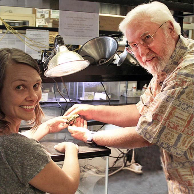 Two individuals in a laboratory setting, one handing a small turtle to the other, both smiling at the scene.