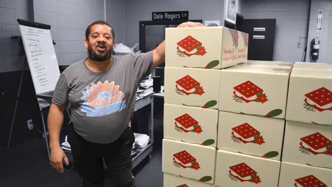 Man posing with his arm up as tall as his head resting it on boxes, the background is black and white.