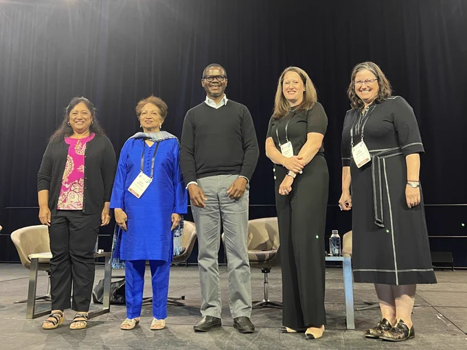 Five speakers are smiling on a stage, with chairs in the background.