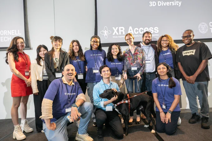 Group shot of XR Access chairs and volunteers onstage at the end of the Symposium. 13 people pose in two rows, along with Shiri’s guide dog Jarvis.
