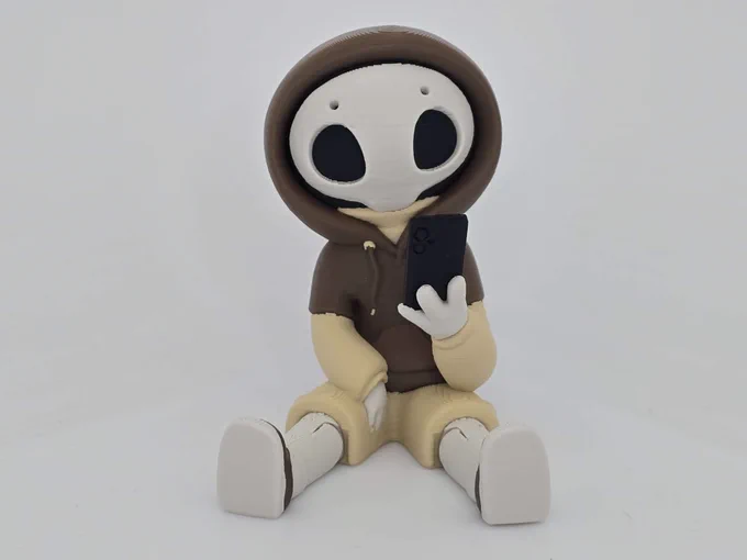 Mocha Hazy figure in brown and cream, seated with legs out, holding a phone and staring ahead on a white backdrop.