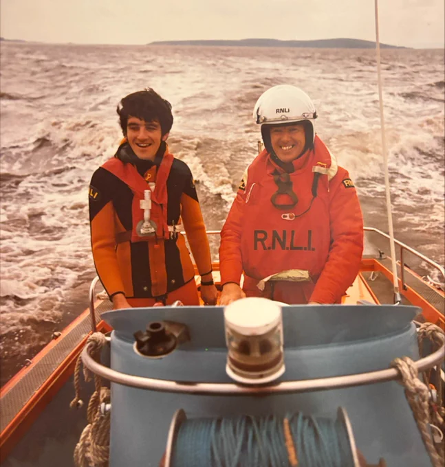 Two individuals in RNLI (Royal National Lifeboat Institution) life jackets smiling on a boat at sea.