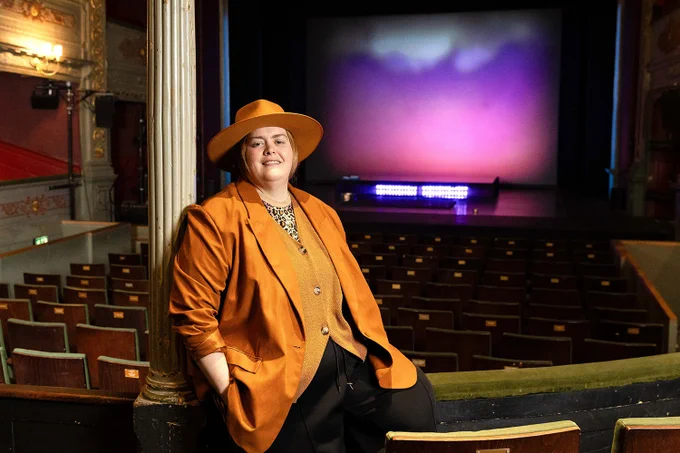 A photo showing a woman sat on a theatre circle front, with the auditorium behind her 