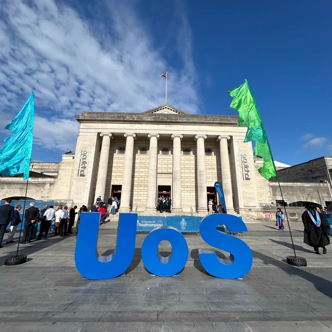 Large blue letters spelling "UoS" are displayed in front of the Guildhall. The sky is bright blue with scattered clouds. 