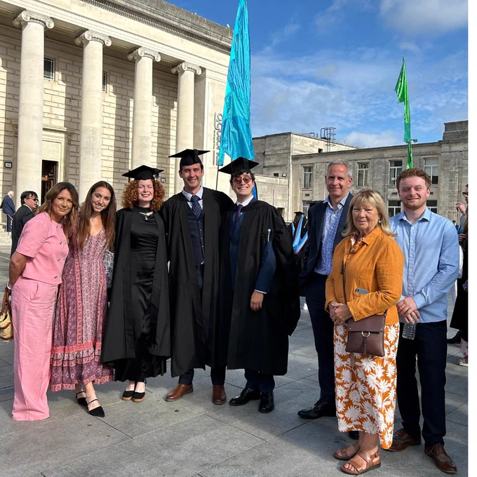 A group stands smiling outside the Guildhall, under a clear blue sky. In the center, three people in graduation gowns and caps with their family standing next to them.