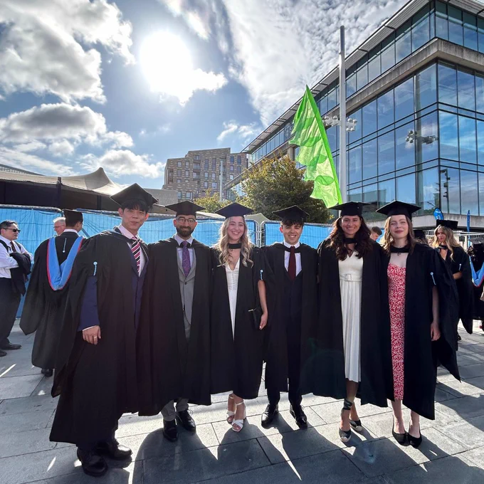 A group of six graduates in caps and gowns pose outdoors under a bright, partly cloudy sky. Behind them is a modern building with large glass windows.