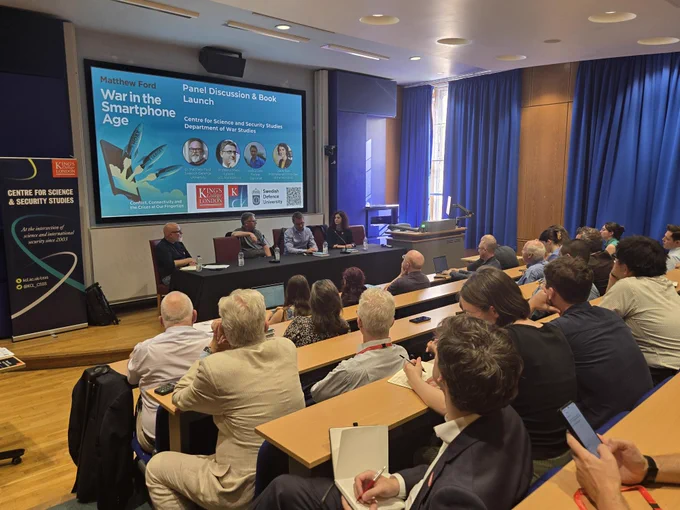 Event photo showing a panel table at the front with speakers, and an audience seated and listening attentively
