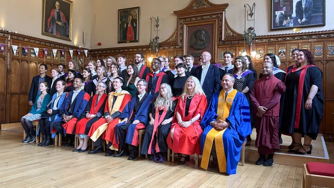 Colour photo of graduating students and staff members stood and standing on the Rainy Hall stage. They are smiling towards a camera that is not visible in the photo.