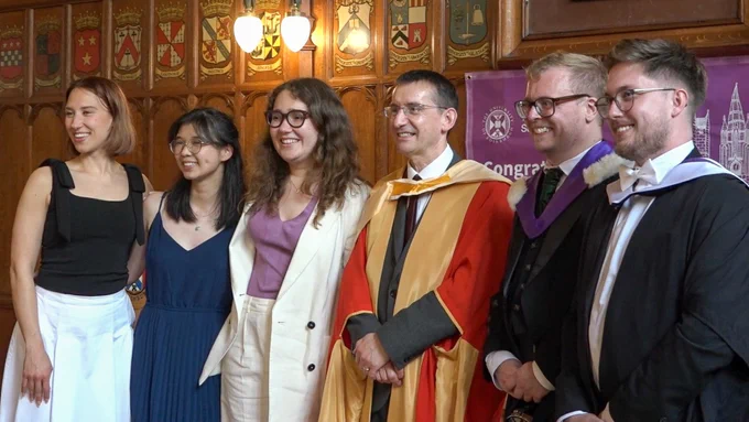 Colour photo of a group of people stood smiling together for a photo. They are looking at a camera off screen.
