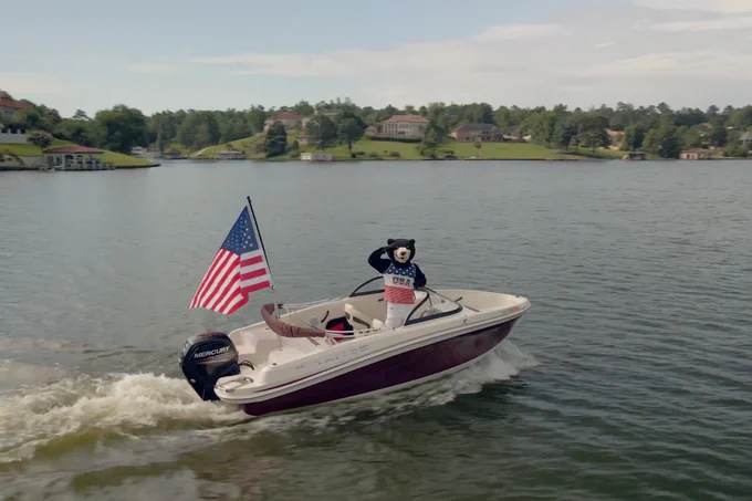 A motorboat featuring a large American flag and a bear mascot dressed as a captain speeds across a lake with houses and greenery in the background.