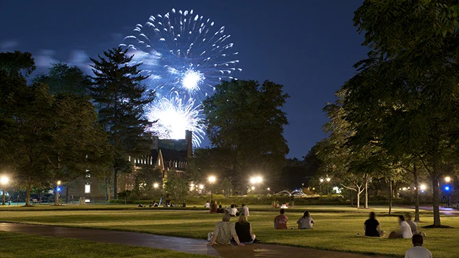 Students sit on the Green watching fireworks over a campus building.