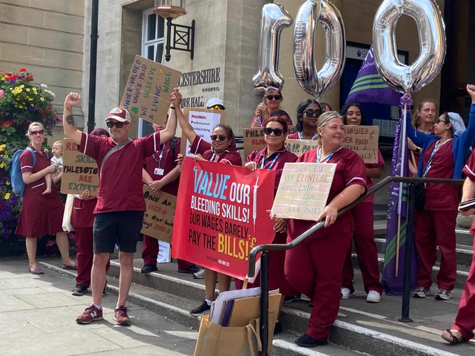 Phlebotomists outside Gloucester Shire Hall with 100 spelt out in helium balloons. They're holding placards banners.