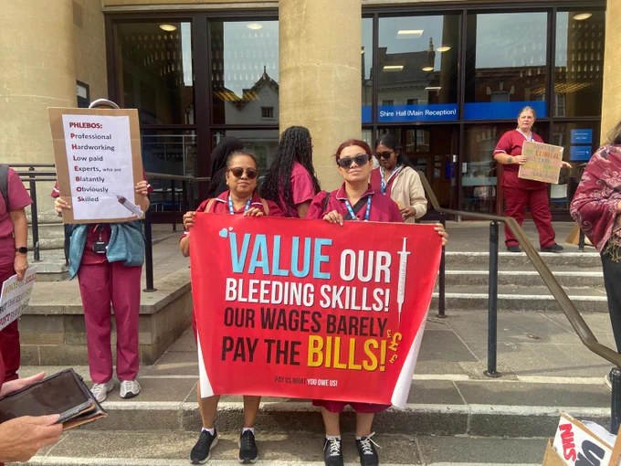 Phlebotomists outside Gloucester Shire Hall with a banner that reads 'value our bleeding skills, our wages barely pay the bills.