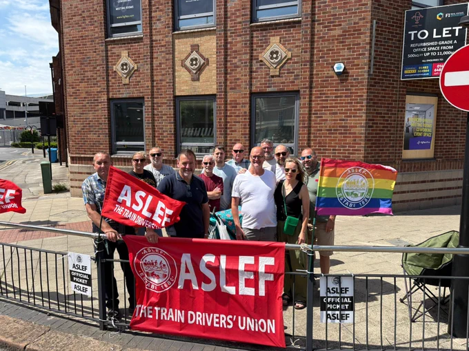 ASLEF Members standing on a picket line in the sun with ASLEF flags and an official picket sign displayed