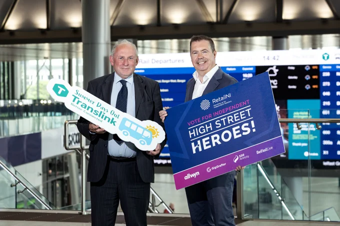 Picture of 2 males, standing in Belfast Grand Central Station. One is holding a photo prop of "Summer Days Out with Translink" and another is holding a photo prop for "Vote for Independent High Street Heroes!"