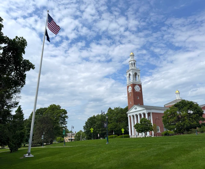 An American flag flying on a pole next to a large grassy area with a view of Ira Allen Chapel, featuring its prominent white facade and golden dome under a blue sky with scattered clouds.
