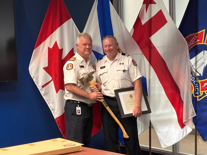 Chief Ken Stuebing presents Division Chief Vince Conrad with his ceremonial retirement axe. They are both standing in front of flags in the trailblazers boardroom. 