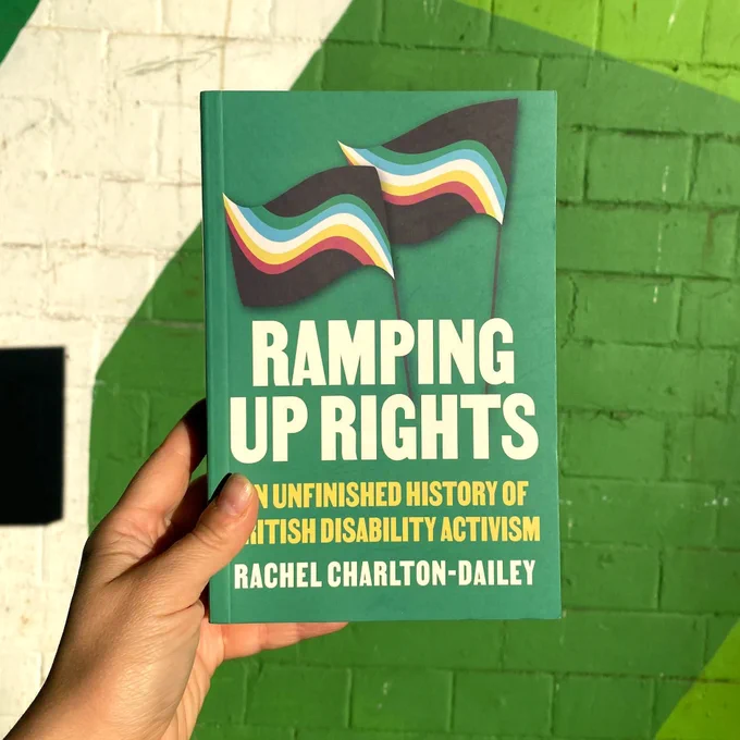 Close-up of a hand holding the book “Ramping Up Rights: An Unfinished History of British Disability Activism” by Rachel Charlton-Dailey. The cover is green with two waving Disability Pride flags and bold white and yellow text. The background is a painted brick wall in shades of green and white.